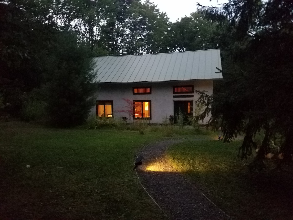 The exterior of the earth home is illuminated during twilight, showcasing large windows that reflect a warm glow. A winding path leads towards the entrance, framed by lush greenery and trees, emphasizing the rustic, natural setting.