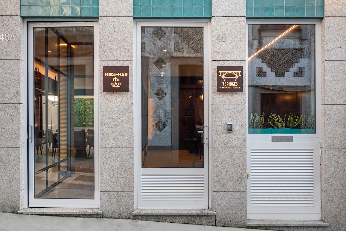 The building exterior features a modern façade with large glass doors leading to two establishments. The entrance is framed by decorative tiles in a subtle green hue, while the adjacent signage identifies the restaurant and hotel. Lush greenery is visible through the windows.