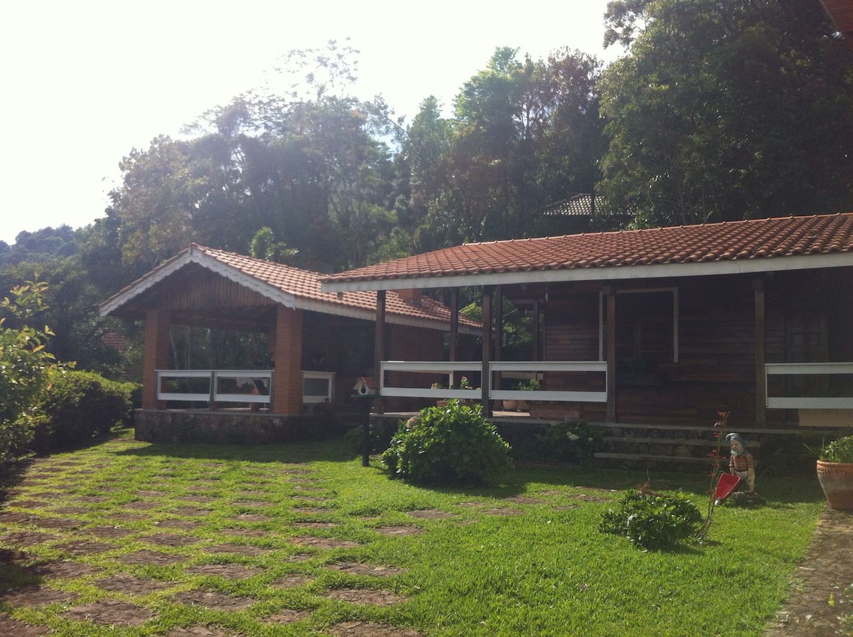 A charming cabin is surrounded by lush greenery, featuring a wooden structure with a sloped roof. An open patio area extends to the side, inviting outdoor relaxation. The neatly arranged stone pathway leads through the vibrant grass, creating a welcoming entrance to the tranquil setting.