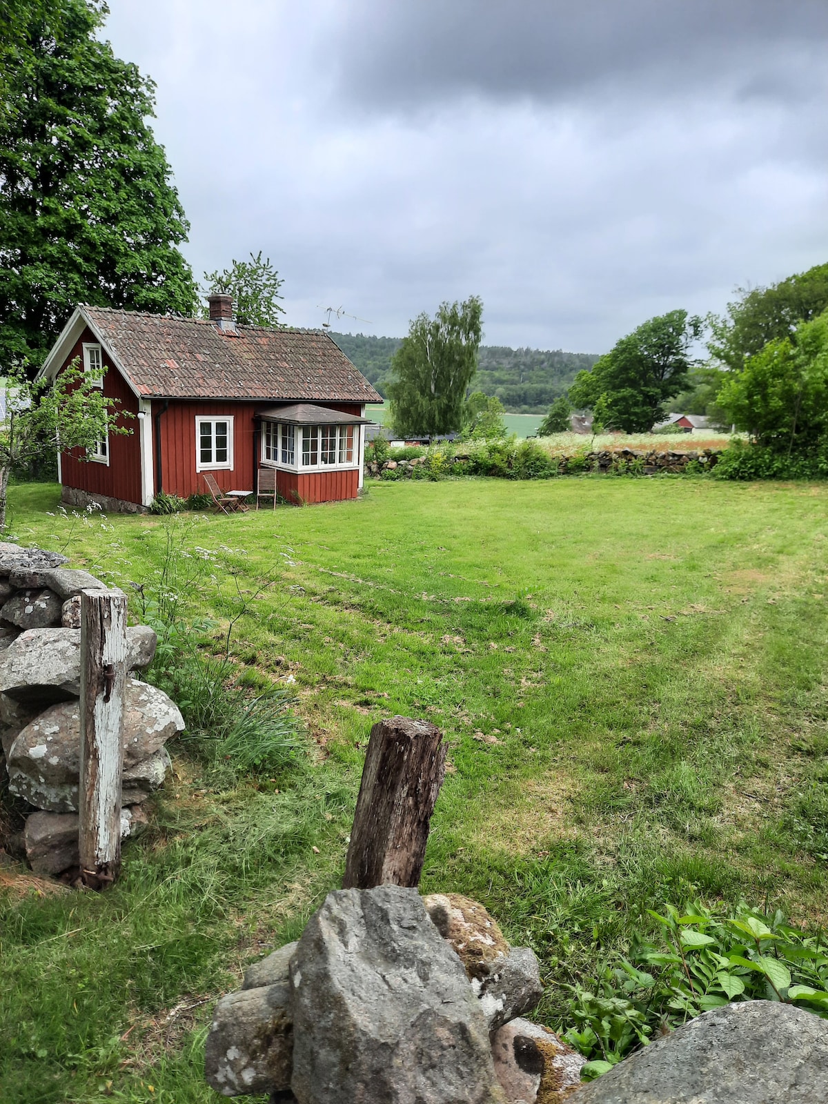 A charming red cottage is surrounded by a lush green lawn and trees, with a stone wall framing the property. The cottage features a glassed-in porch and a sloped roof, set against a cloudy sky and open landscape in the background.
