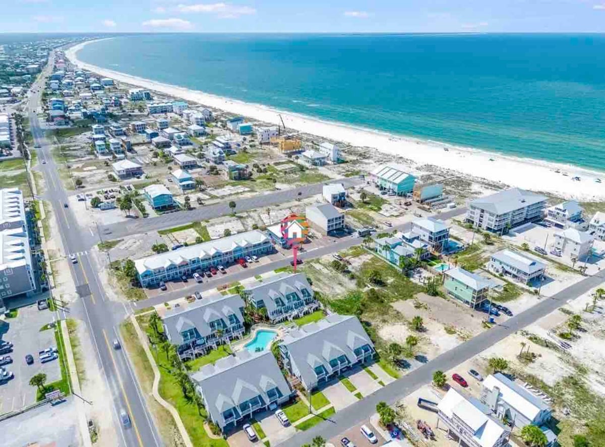 An aerial view captures the townhouse's coastal location, with sandy beaches stretching along the shoreline. Surrounding areas show a mix of residences and local businesses, while the turquoise waters of the sea contrast with the light-colored sand, inviting beach activities.