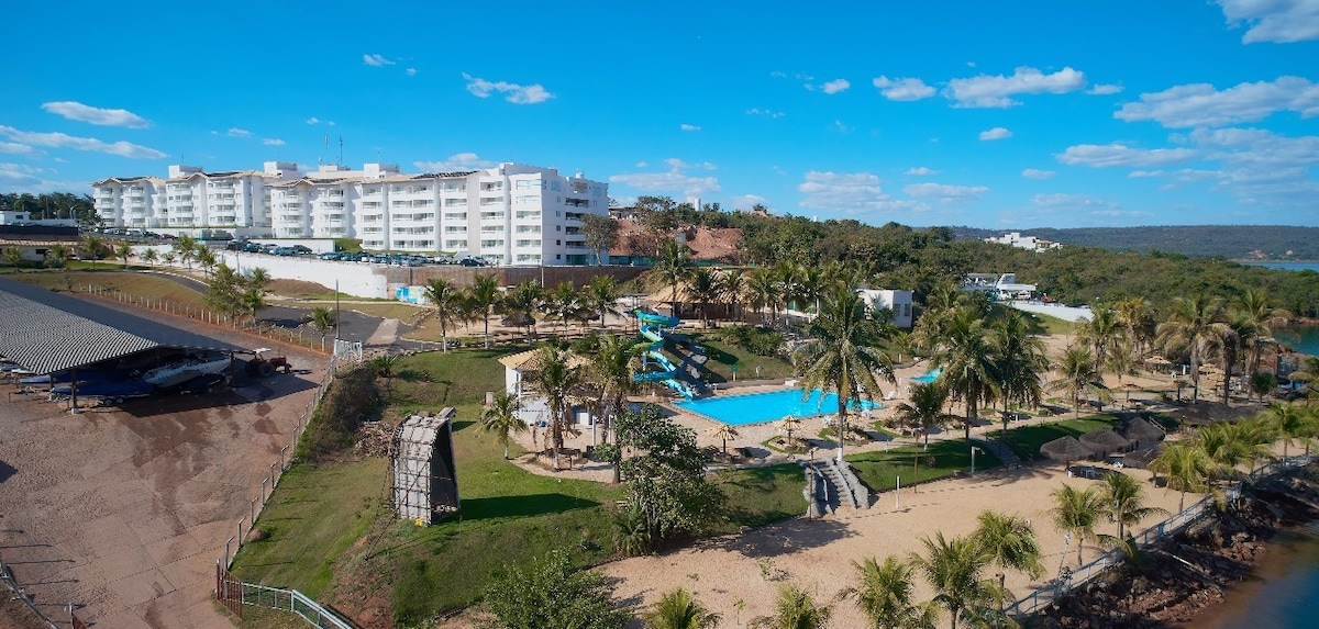 An aerial view captures the resort layout, featuring a large aquatics area with multiple water slides and a spacious pool. Lush palm trees adorn the landscape, providing a tropical feel. The modern white buildings are set against a scenic backdrop of blue skies and rolling hills.