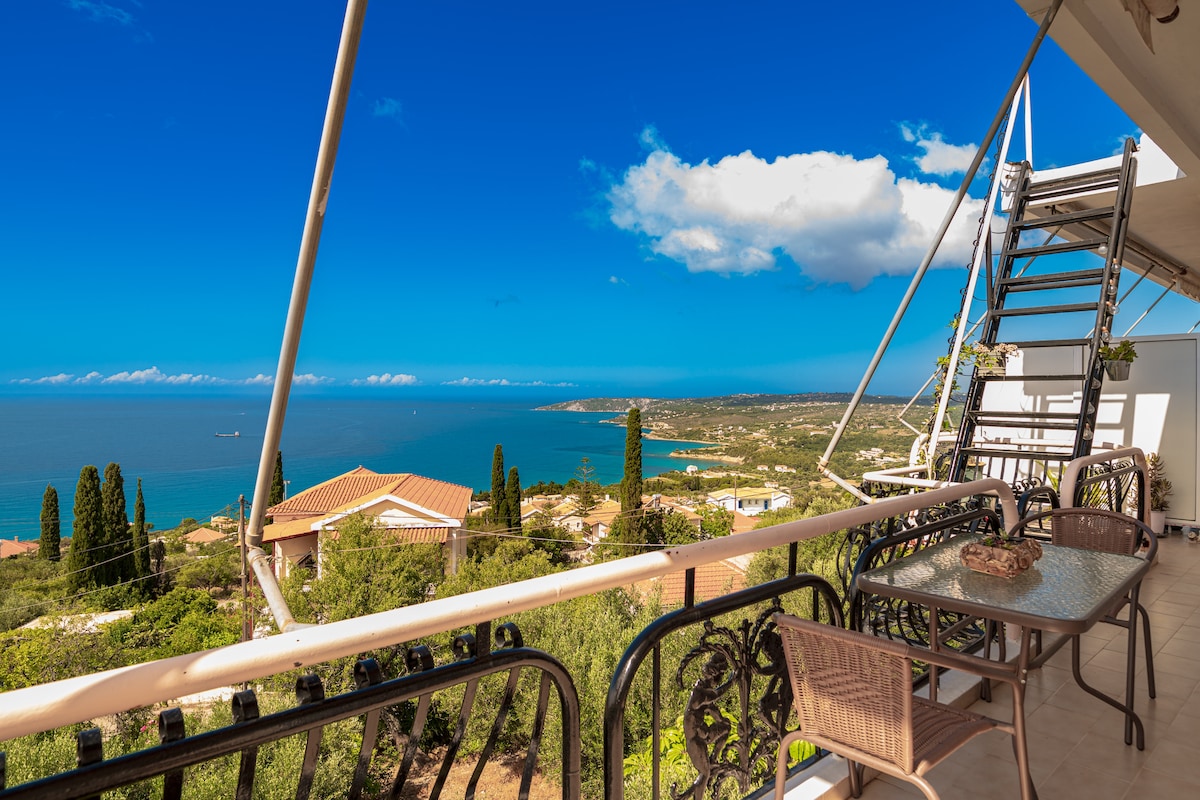 A balcony area provides breathtaking views of the sea and mountains, framed by greenery. A small round table with four chairs is positioned beside the balcony railing, allowing for outdoor relaxation. The sky is clear with scattered clouds above the distant coastline.