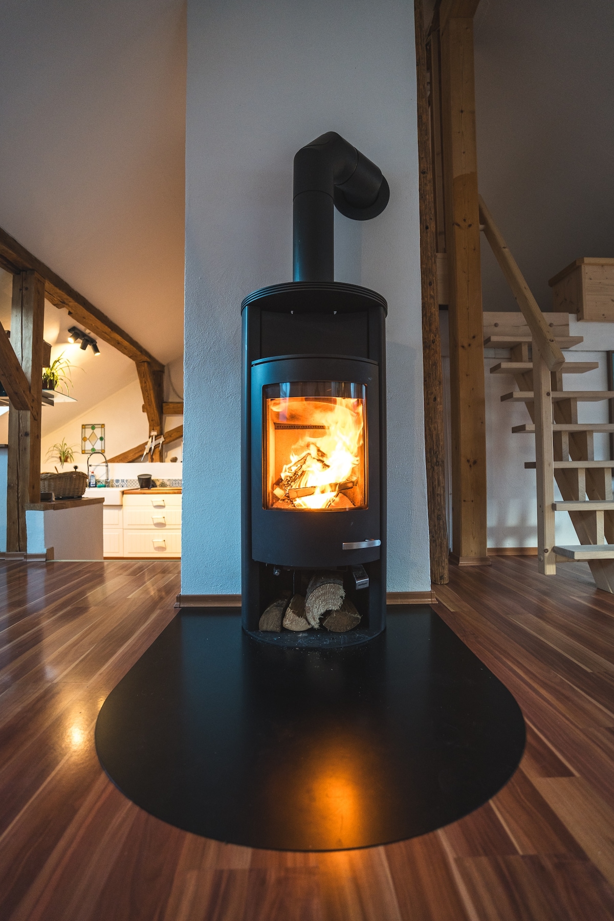 A modern wood stove stands against a wooden beam wall, with flames gently flickering inside. The stove is placed on a round black base, complementing the polished wooden floor that reflects light. The background reveals a glimpse of the kitchen area and staircase.