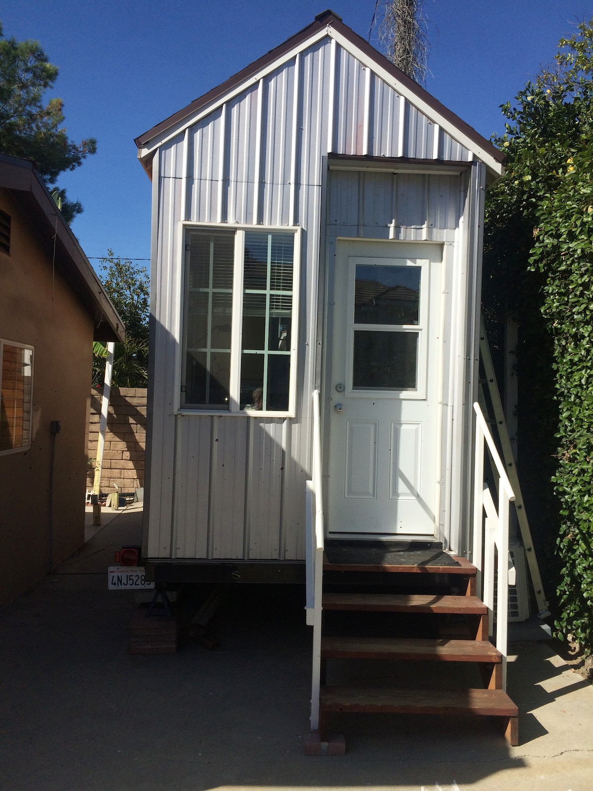 A compact tiny house features a metal exterior with vertical panels. A white front door is flanked by rectangular windows, allowing natural light to enter. Steps lead up to the entrance, surrounded by greenery and adjacent to a neighboring structure.