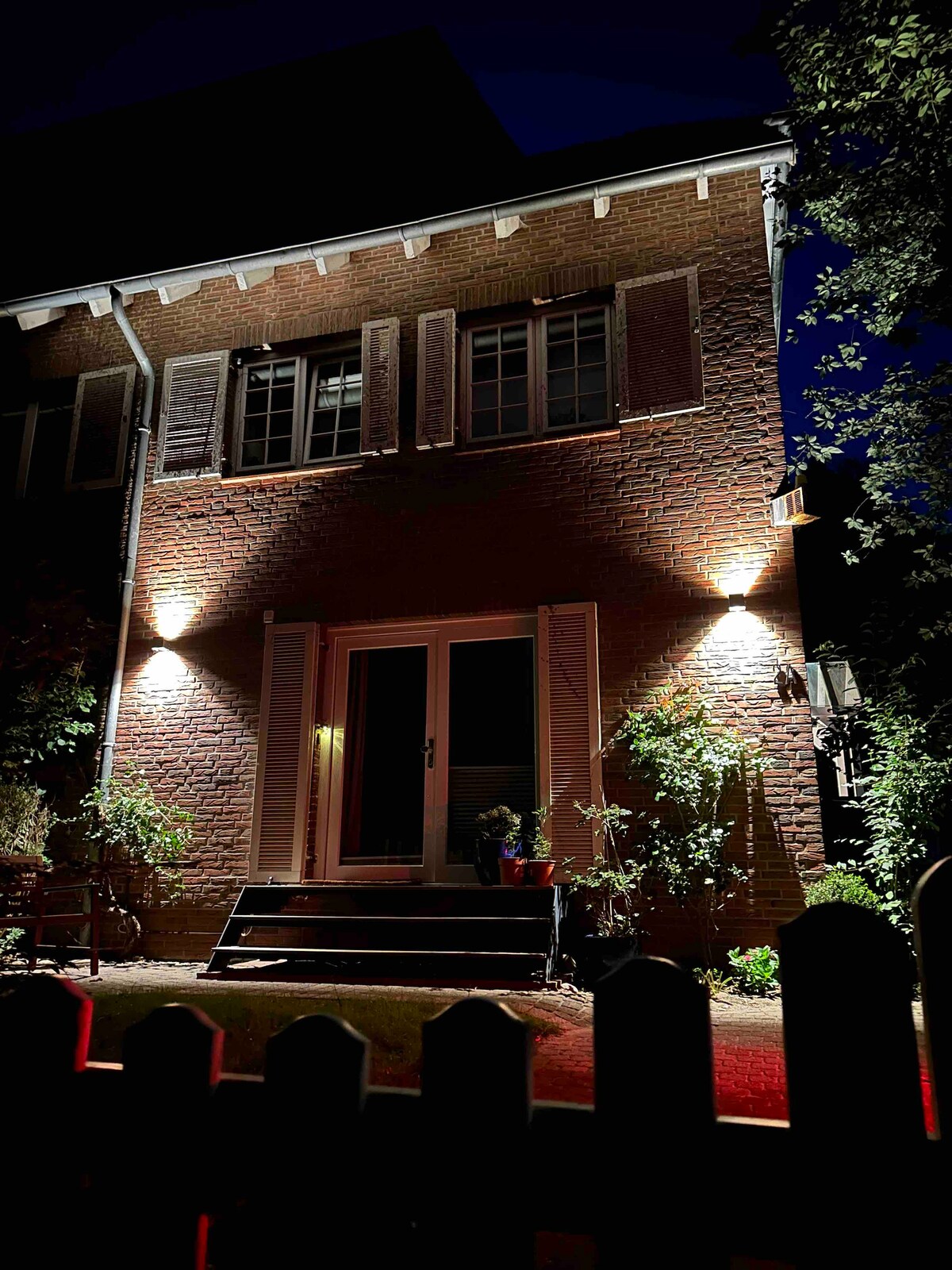 The two-story brick house is illuminated at night, highlighting its elegant shutters and welcoming entryway. Soft light casts a warm glow over the garden area, enhancing the natural surroundings. A set of steps leads to the entrance, framed by greenery and decorative details.