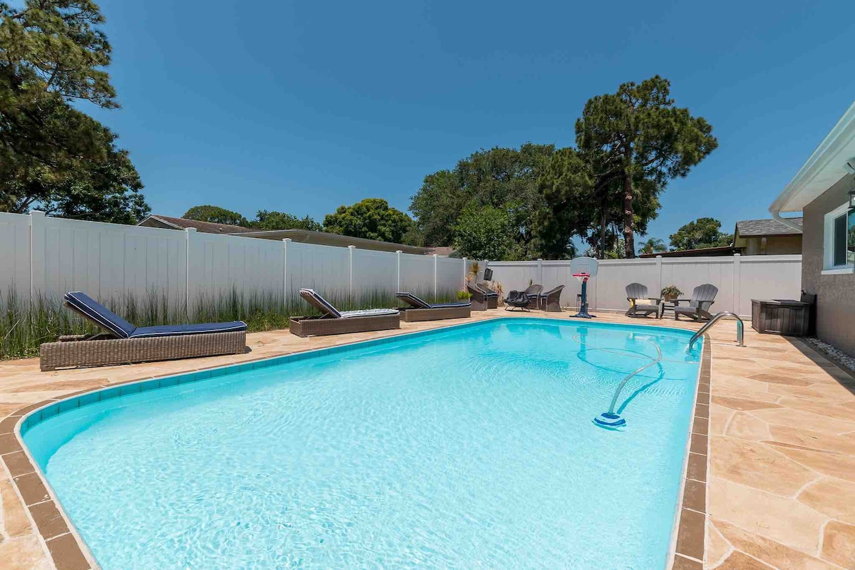 A spacious heated pool is surrounded by lounge chairs and patio furniture, set against a backdrop of green trees and a white privacy fence. Sunlight reflects off the clear water, inviting relaxation and enjoyment in a serene outdoor space.