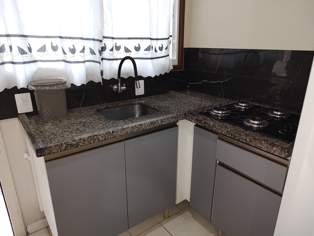 The kitchen features modern cabinetry with a combination of gray and white finishes. A granite countertop offers ample workspace, while a stainless steel sink and a four-burner gas cooktop are included. Natural light enters through a window adorned with a simple curtain.