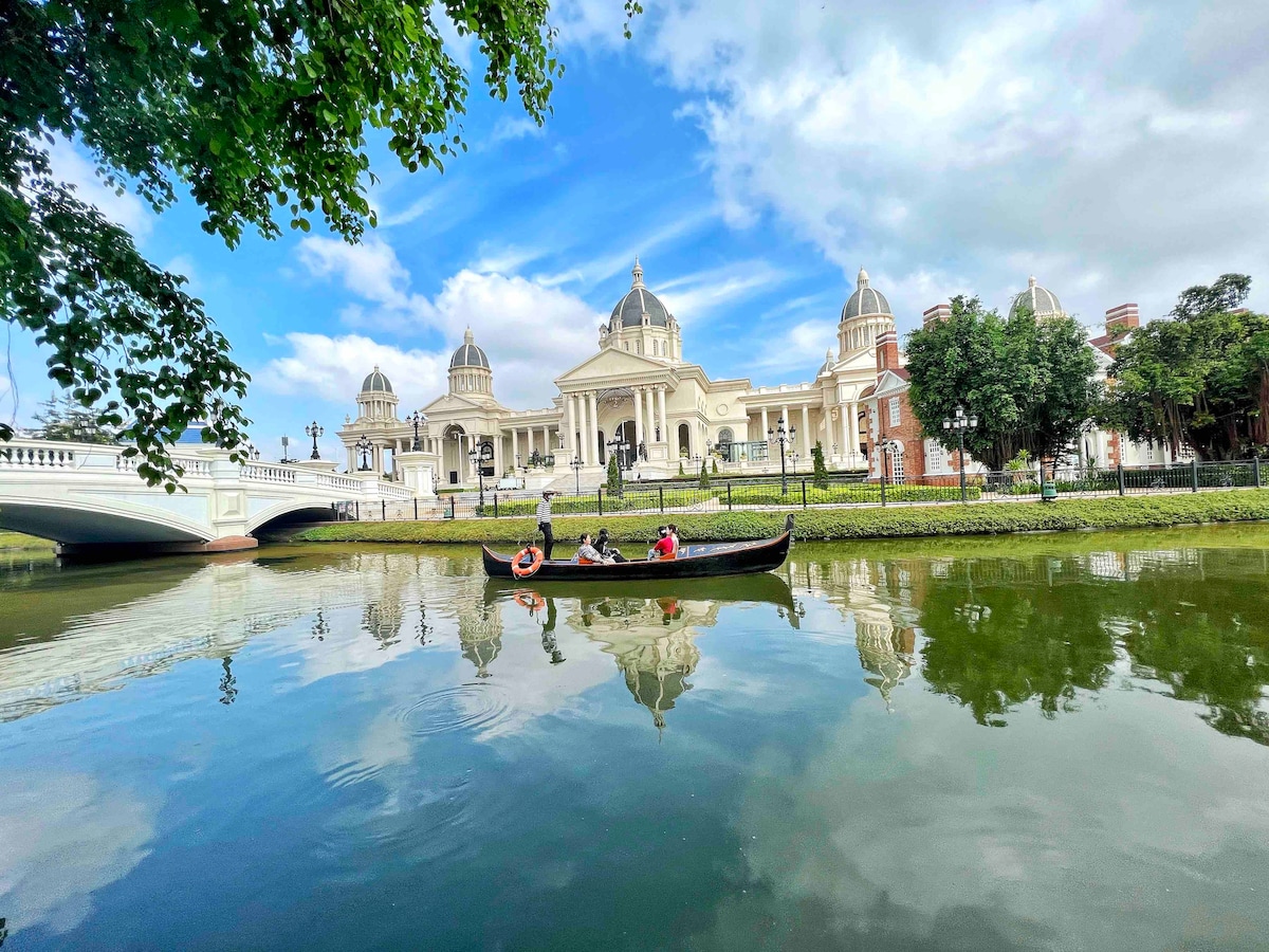 A serene view captures a traditional boat gliding on calm waters, reflecting a grand building with domed roofs in the background. Lush greenery frames the scene, while a bridge spans over the water, enhancing the tranquil ambiance of the area.
