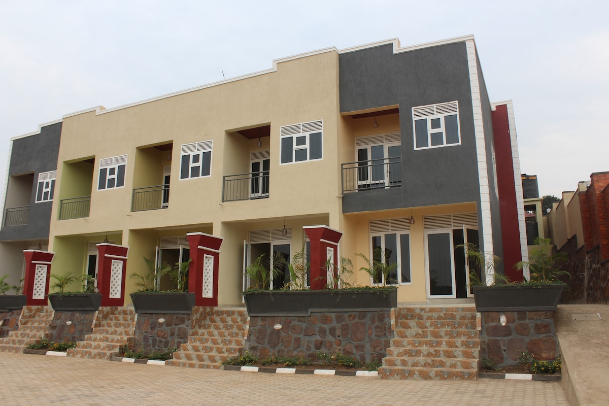 A modern villa structure is displayed, featuring two levels with multiple balconies. Each unit showcases large windows and colorful exteriors, emphasizing a blend of neutral and bold tones. Well-maintained landscaping is visible in front of the building, with potted plants enhancing the entrance.