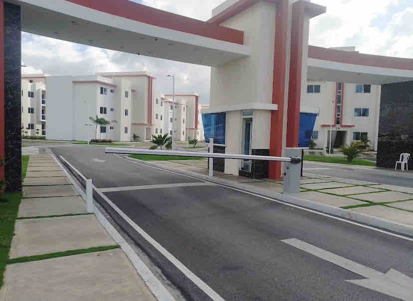 The entrance of a secure residential complex is shown, featuring a gated access point with a barrier. The surrounding area includes neatly arranged pathways and landscaped sections, leading towards the multi-story apartment buildings in the background.