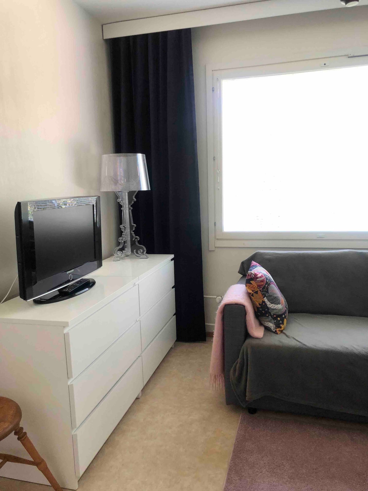 A cozy corner of the living room features a gray sofa adorned with a decorative pillow. A sleek white dresser stands beside a flat-screen TV. Light filters through a partially covered window, complemented by dark curtains.