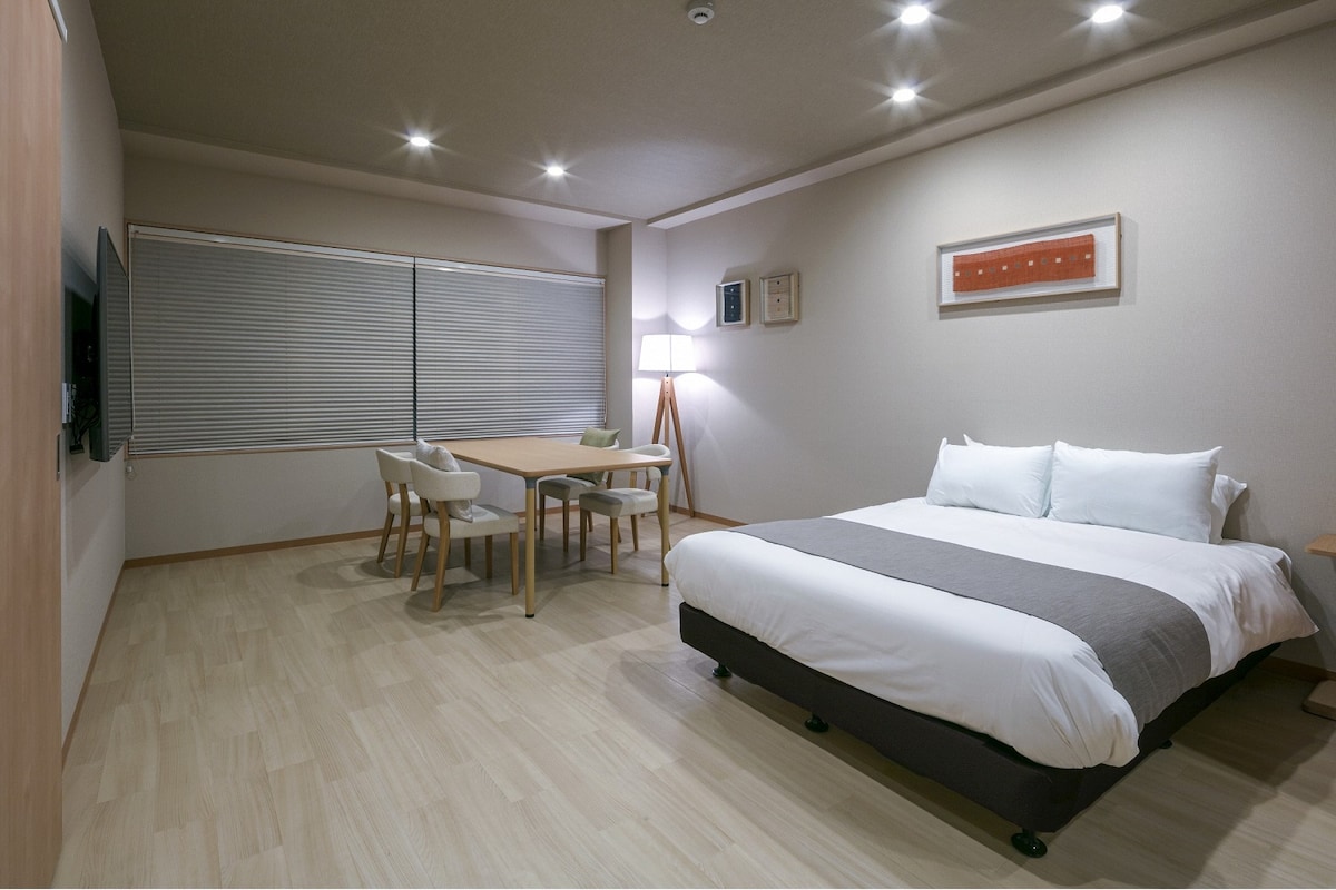 A well-lit room features a spacious bed with a grey runner, wooden furniture, and two chairs. A dining table is positioned near large windows with blinds. A flat-screen TV and ambient lighting enhance the cozy atmosphere.