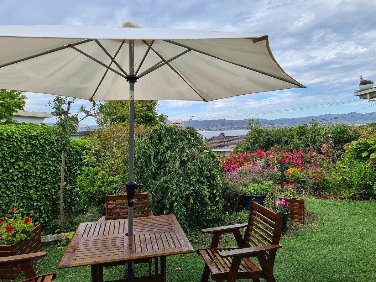 An outdoor dining area features a wooden table with chairs, shaded by a large umbrella. Lush greenery surrounds the space, complemented by colorful flowerbeds. The view beyond includes a serene river and distant hills under a lightly clouded sky.