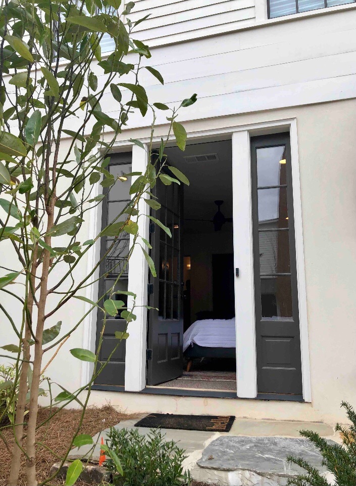 The entrance features double doors painted in a dark hue, flanked by greenery. Through the doors, a glimpse of the bedroom is visible, showcasing a neatly made bed and inviting interior. A doormat lies in front, providing a welcoming touch.