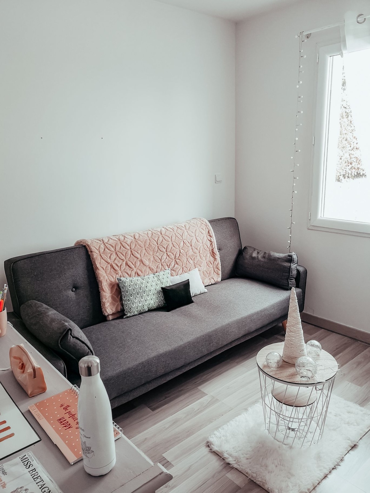 A compact sitting area features a gray sofa with a textured throw and decorative pillows. A small white table is positioned nearby, topped with a minimalistic bottle and a decorative cone. Light enters through a nearby window, highlighting the soft tones of the space.