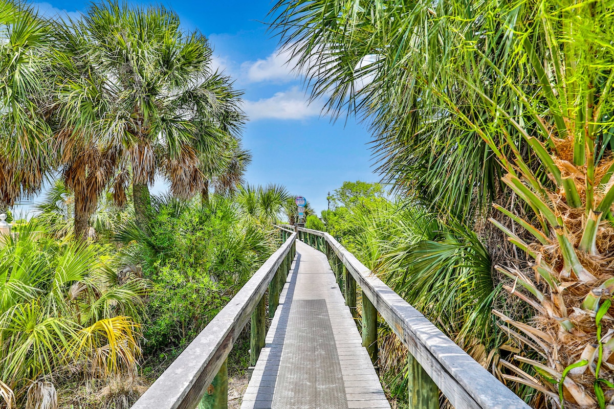 A wooden boardwalk is surrounded by lush tropical vegetation, leading towards a clear blue sky. The pathway is flanked by palm trees and various green shrubs, providing a serene and nature-filled atmosphere.