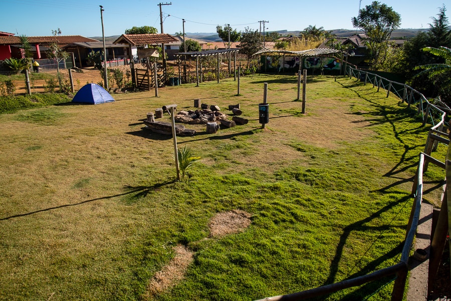 An open grassy area is highlighted, featuring a fire pit surrounded by natural seating made of logs. A tent is pitched on the grass, while rustic structures and trees create a serene backdrop. Fencing and pathways lead to various outdoor spaces.
