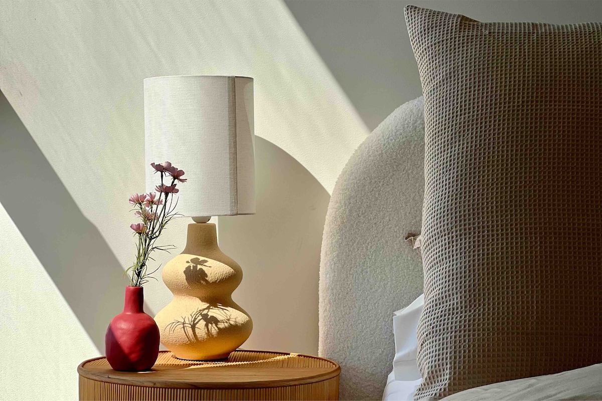 A bedside table features a textured lamp with a beige shade and a yellow base. A small red vase holds delicate flowers. The bed is adorned with a large beige pillow, and soft light casts gentle shadows on the wall.