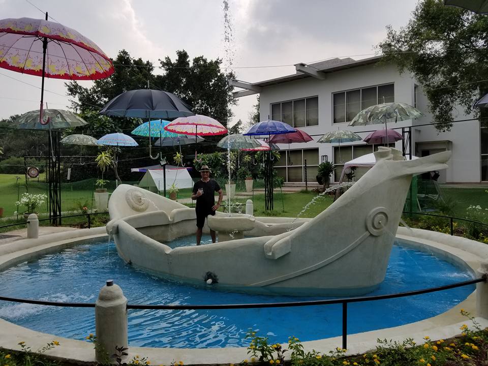 A whimsical pool area features a boat-shaped sculpture surrounded by clear blue water. Colorful umbrellas are displayed overhead, providing shade. Lush greenery and a well-manicured garden are visible in the background, creating a relaxing atmosphere.