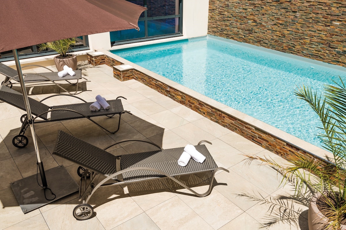 A seasonal outdoor pool glistens under the sun, surrounded by sleek stone walls. Relaxing lounge chairs are positioned nearby with neatly rolled towels. An umbrella provides shade, while potted plants enhance the tranquil ambiance.