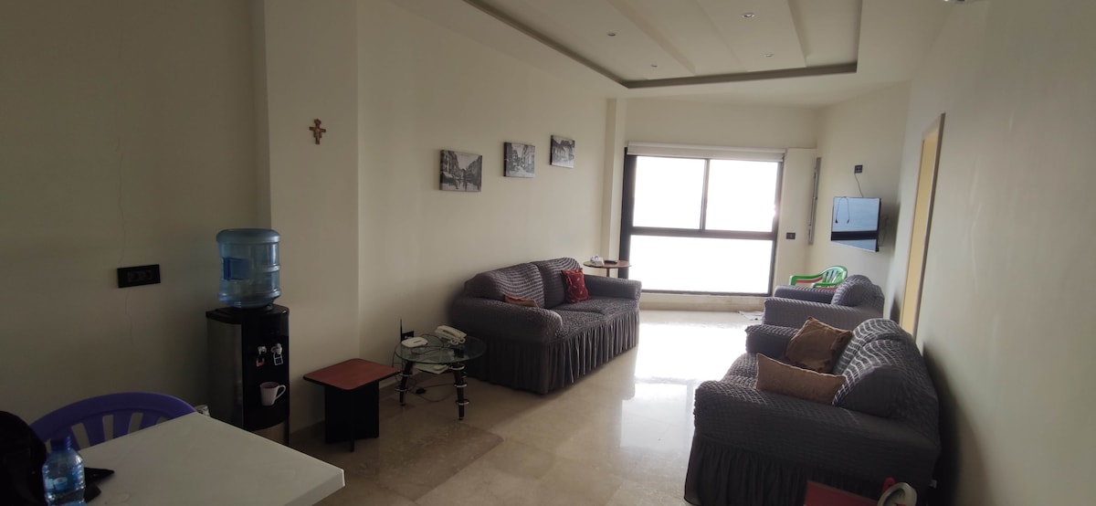 A living area is presented with two spacious sofas and a glass coffee table. Light enters from a large window, complementing the neutral wall color. A water dispenser and a TV are visible, while wall art adds simple visual interest to the space.