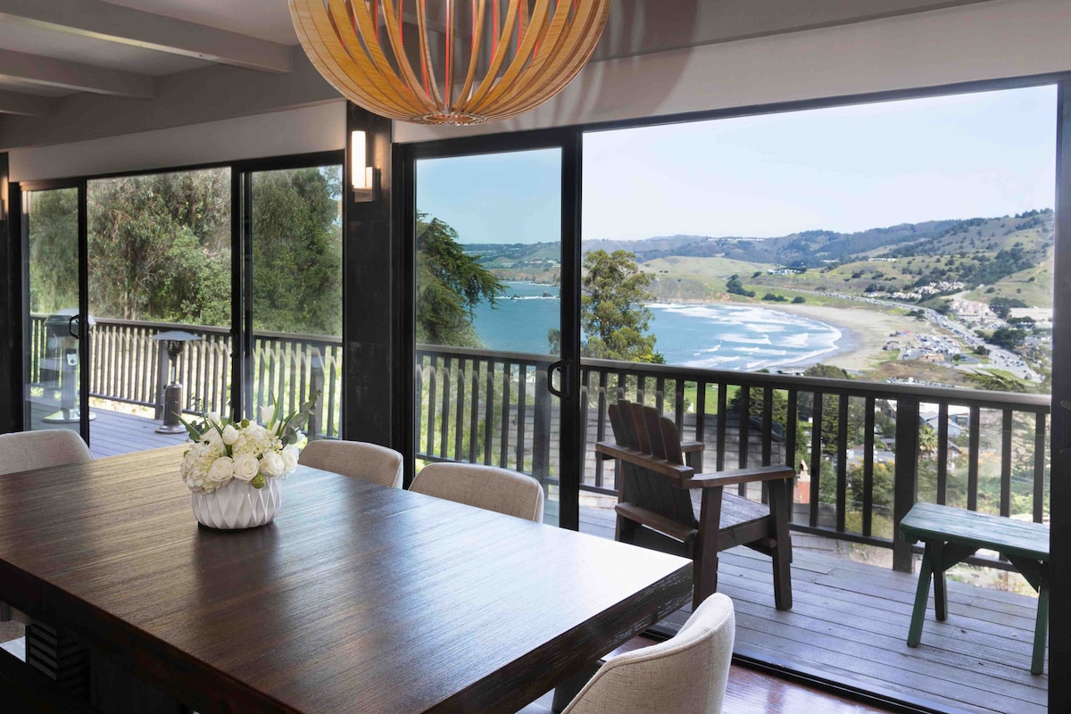 A dining area is presented with a large wooden table surrounded by light-colored chairs. Floor-to-ceiling windows offer unobstructed ocean views, while a decorative light fixture hangs above. A cozy seating area with wooden chairs is situated on the deck outside.