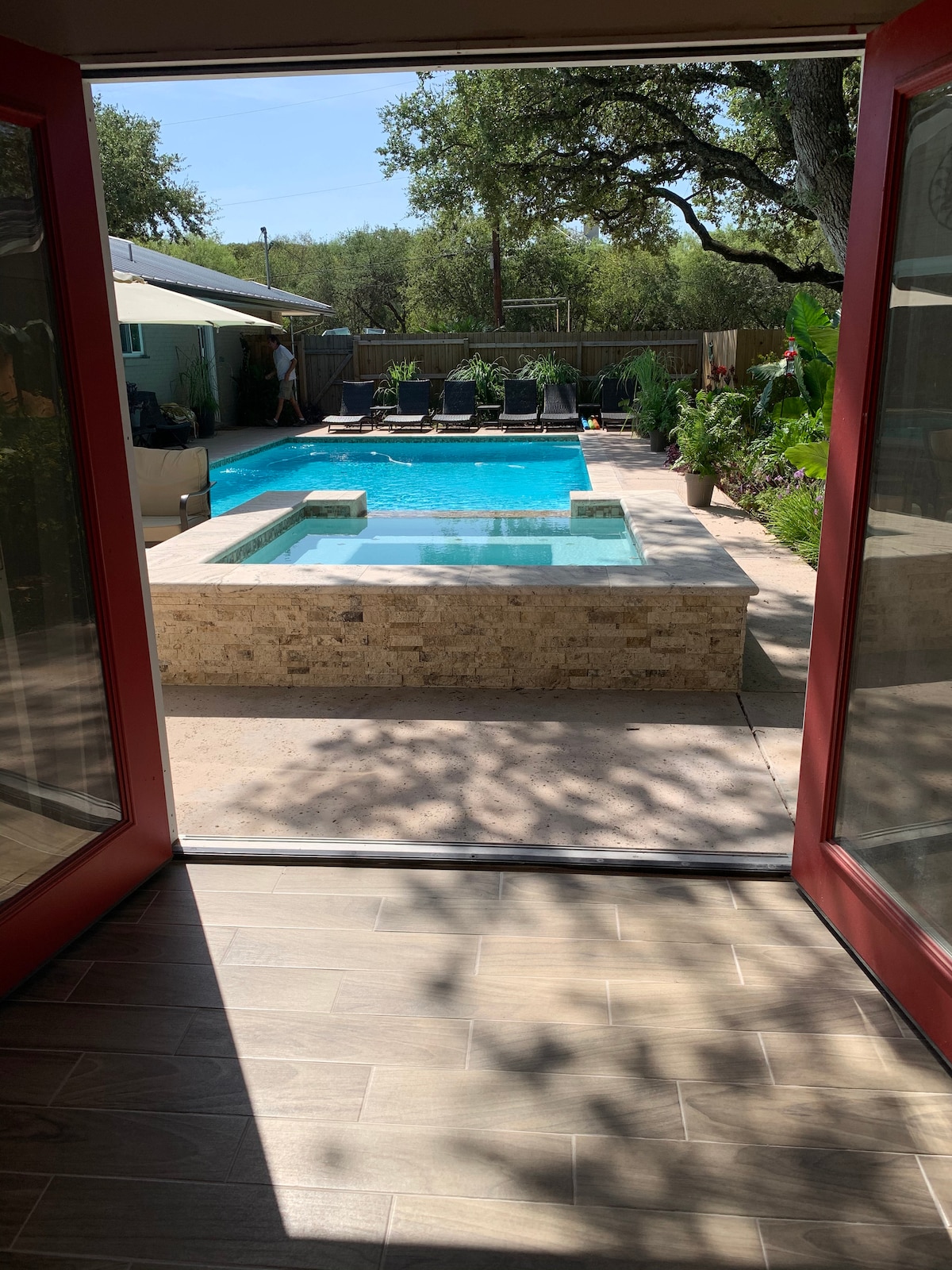 The view from the open door showcases a serene outdoor space featuring a crystal-clear pool and a hot tub, surrounded by lush greenery and lounge chairs. Sunlight filters through trees, creating patterns on the patio floor.