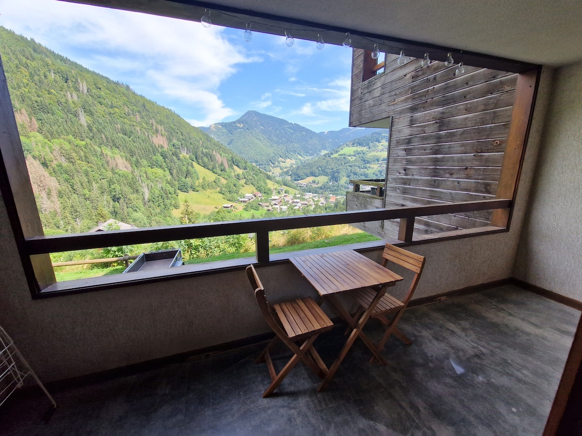 A balcony area features a wooden table with two foldable chairs, providing a view of the valley and surrounding mountains. Large windows allow natural light to fill the space, and the rustic wooden structure adds to the peaceful atmosphere.