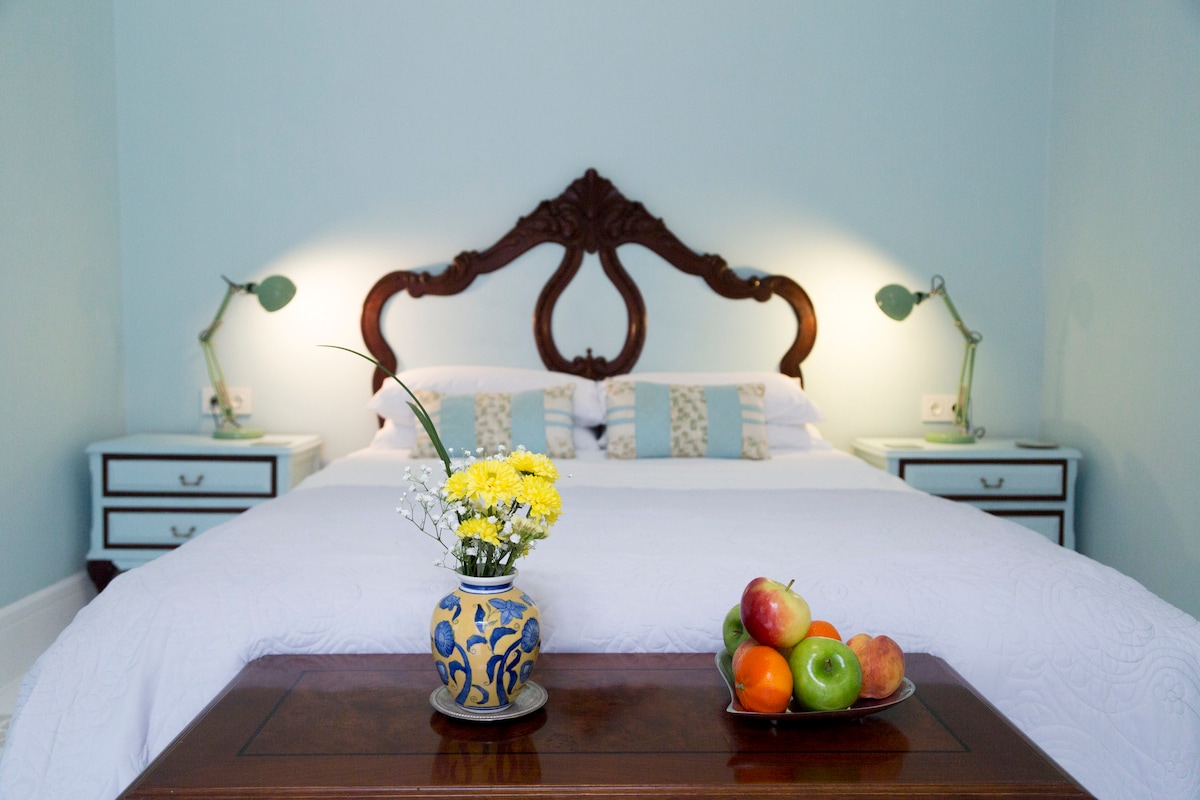 A serene bedroom features a spacious bed with a decorative headboard, flanked by two bedside tables with green lamps. A wooden coffee table holds a vase of fresh flowers and a bowl of assorted fruit. Soft light enhances the calming atmosphere.