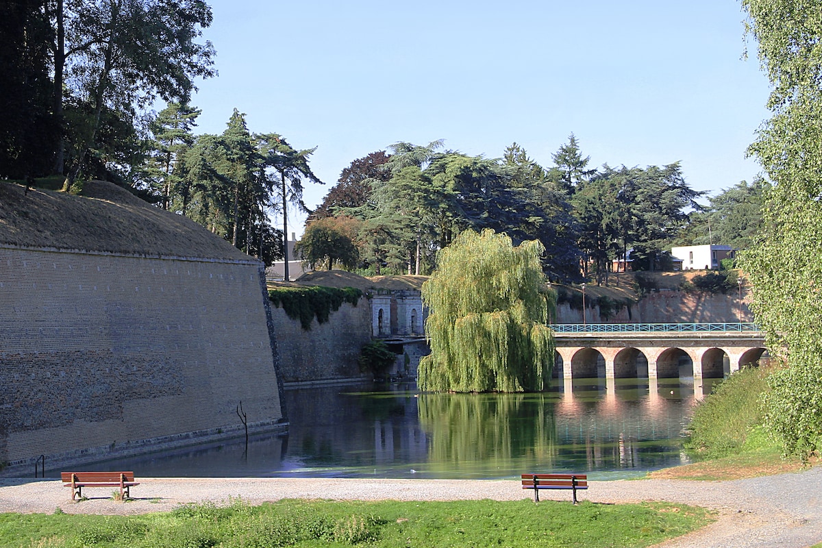 The image depicts a serene outdoor landscape featuring a calm water body surrounded by greenery. Historical stone walls can be seen on one side, alongside a bridge arching over the water. Several benches are positioned near the water's edge, inviting relaxation.