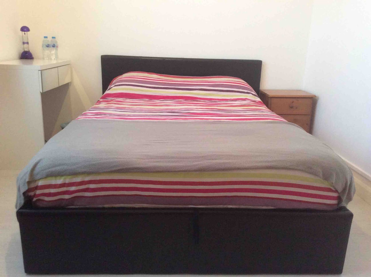 A spacious bed is centered in the image, featuring a striped duvet in shades of red, pink, and yellow. A bedside table is positioned beside the bed, with a small lamp and two bottles of water displayed on a minimalist desk nearby.