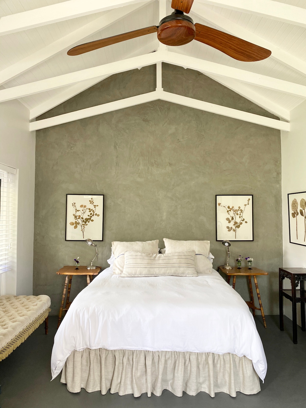 A cozy sleeping area features a queen size bed draped with white bedding and accented by two decorative pillows. The walls are finished in a soft gray, complemented by art pieces depicting botanical designs. Natural light enters through a window, while a ceiling fan circulates air above.