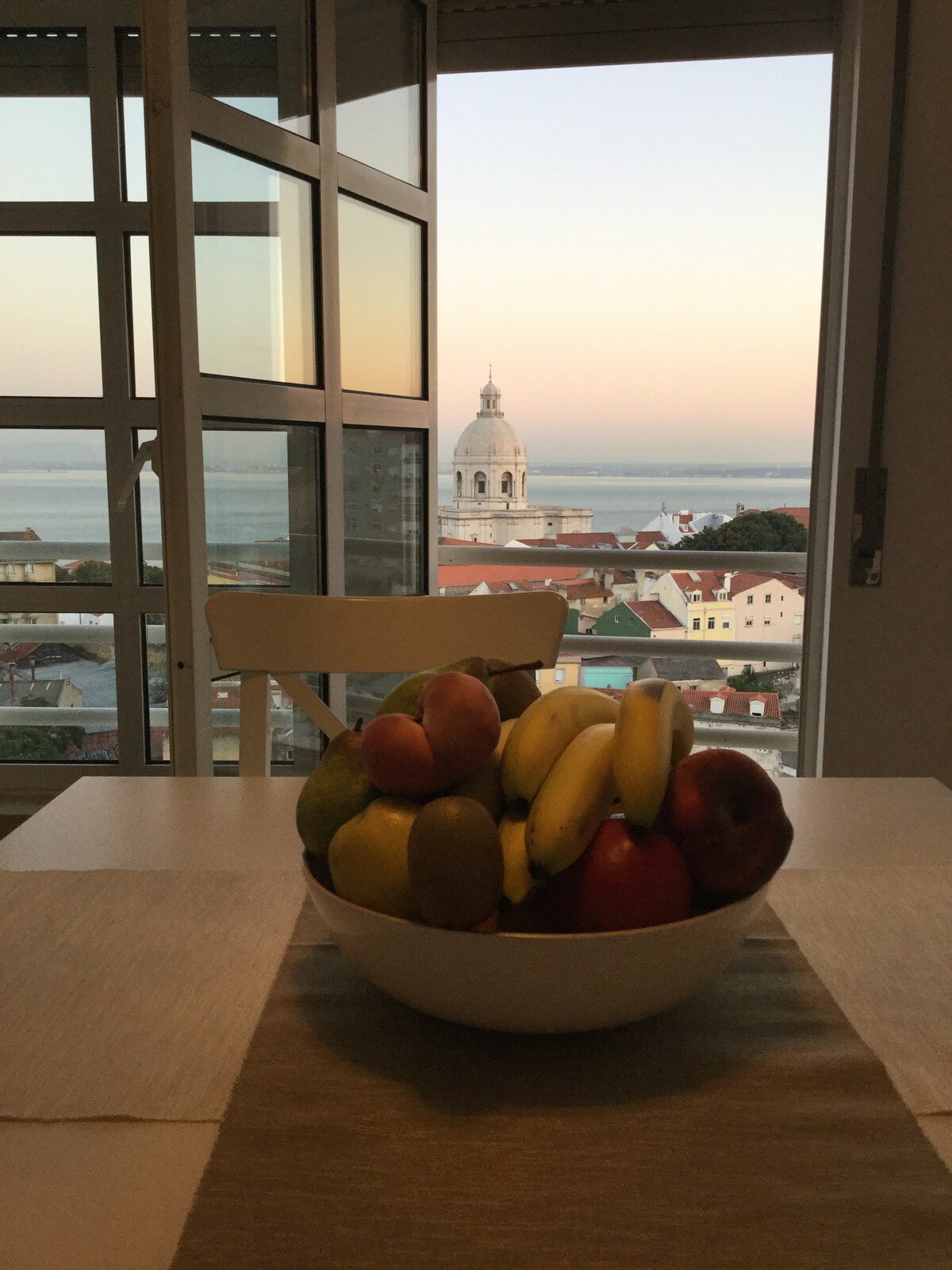 A bowl of assorted fruits, including bananas, apples, and oranges, is centered on a dining table. In the background, expansive windows frame a view of the sunset over a cityscape, with a bell tower and the water subtly visible.