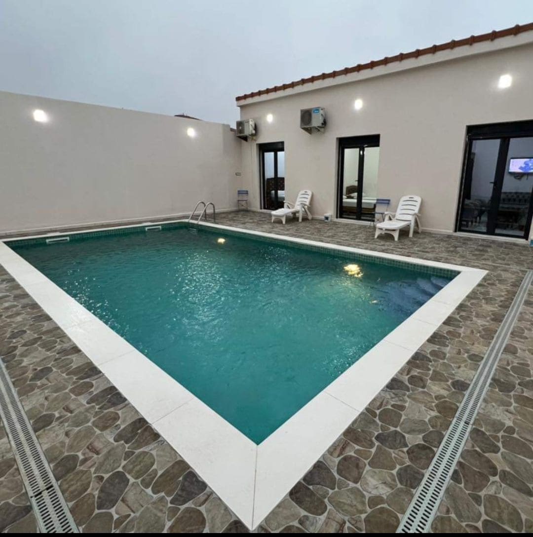 A rectangular pool with a clear, blue surface is surrounded by a stone-tiled area. Lounge chairs are positioned along the poolside. Large glass doors lead to an indoor space, enhancing the connection between indoor and outdoor areas.