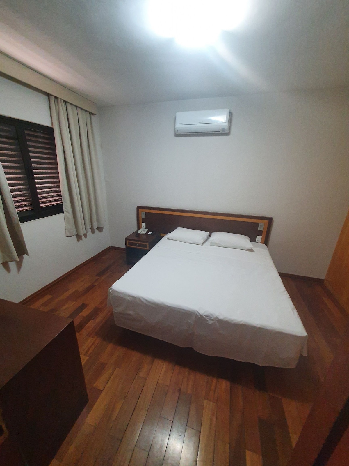 The bedroom features a bed with white bedding positioned against a wooden headboard. Light wooden flooring enhances the space, which is illuminated by ceiling lighting. A bedside table is placed next to the bed, and window coverings allow for light control.