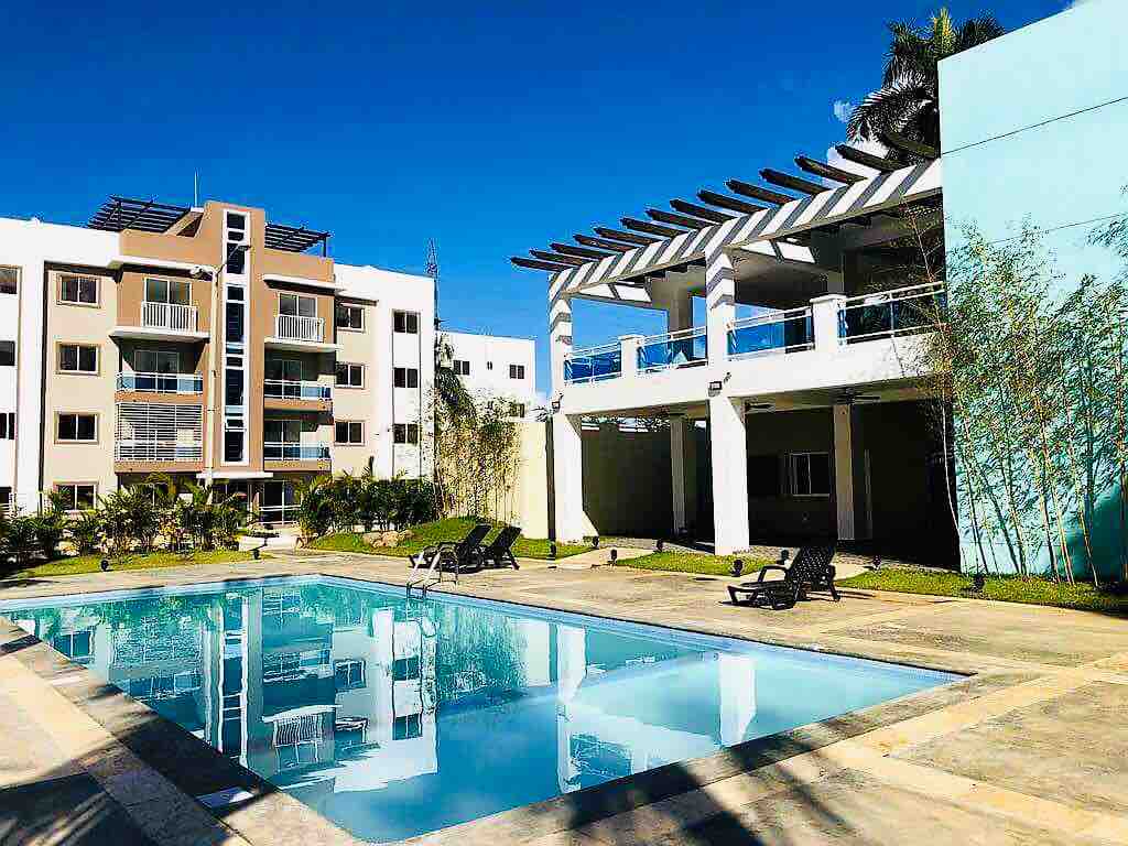 An outdoor pool area features a clear water pool surrounded by lounge chairs. Adjacent structures showcase modern architecture with large windows and balconies, providing an open atmosphere. Lush greenery enhances the setting, contributing to a relaxing environment.