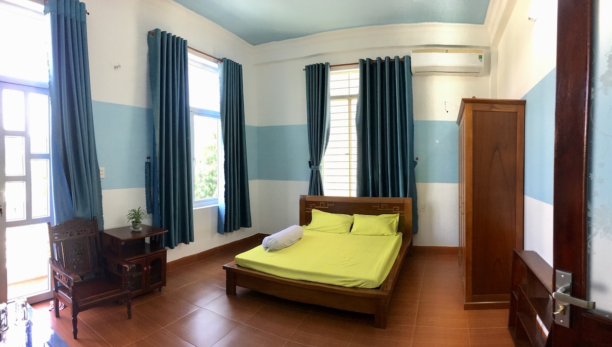 A bright bedroom features light blue and white walls, complemented by tall windows adorned with dark curtains. The spacious bed is covered with a vibrant yellow blanket. A wooden wardrobe and a small side table with a plant are positioned in the room, enhancing its functionality.