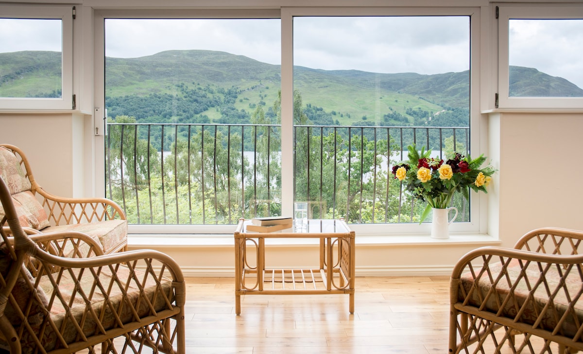 A scenic view of rolling hills is framed by large glass windows, allowing natural light to fill the space. Two comfortable rattan chairs are positioned around a small coffee table, decorated with a vase of fresh flowers, inviting relaxation.
