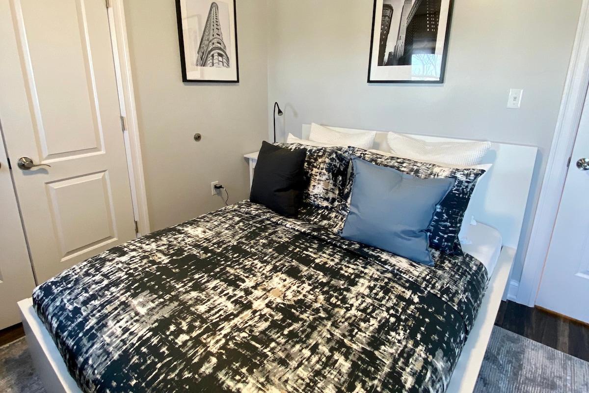 A cozy bedroom is presented with a modern white bed featuring a dark patterned comforter and multiple pillows. Black and white framed photographs adorn the walls, enhancing the space's aesthetic. A bedside lamp is positioned on a matching nightstand, providing a functional and stylish touch.
