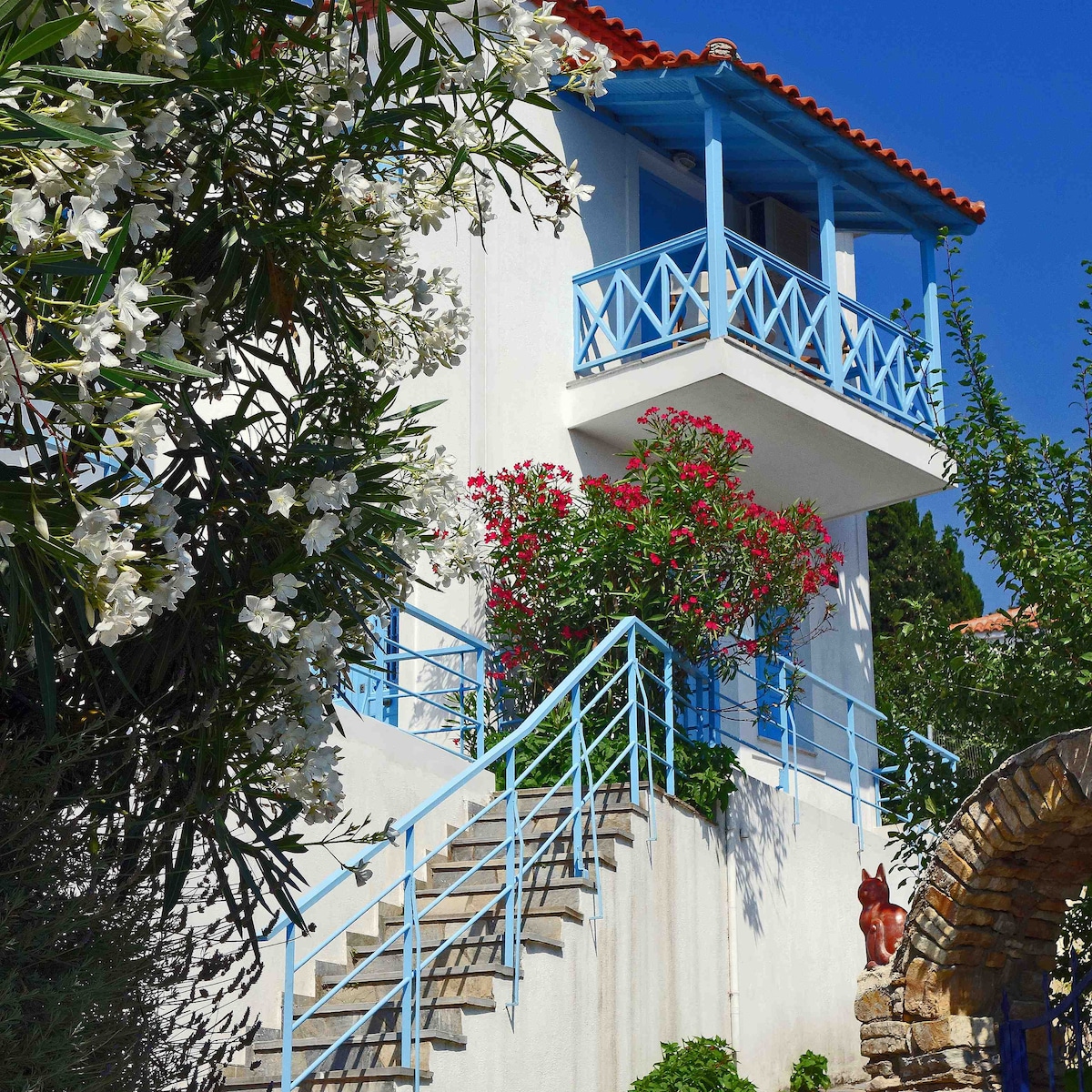 A two-level house is depicted, featuring a white exterior and a red-tiled roof. A blue balcony with railings overlooks vibrant flowering plants below. A curved stone pathway leads towards the entrance, framed by greenery, creating a serene outdoor environment.