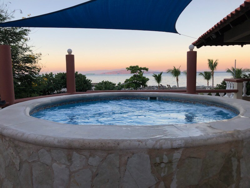 A round jacuzzi is positioned on a stone patio, overlooking the ocean. Its clear water reflects the soft hues of the sunset. Lush greenery and palm trees are visible in the background, enhancing the serene coastal view.