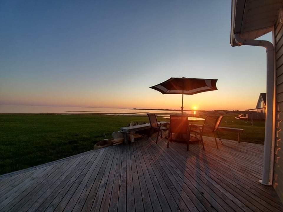 A wooden deck is featured with a dining table and four chairs, alongside a large umbrella providing shade. The sun sets over the calm waters of the Northumberland Strait, creating a serene backdrop of soft hues reflected on the surface.