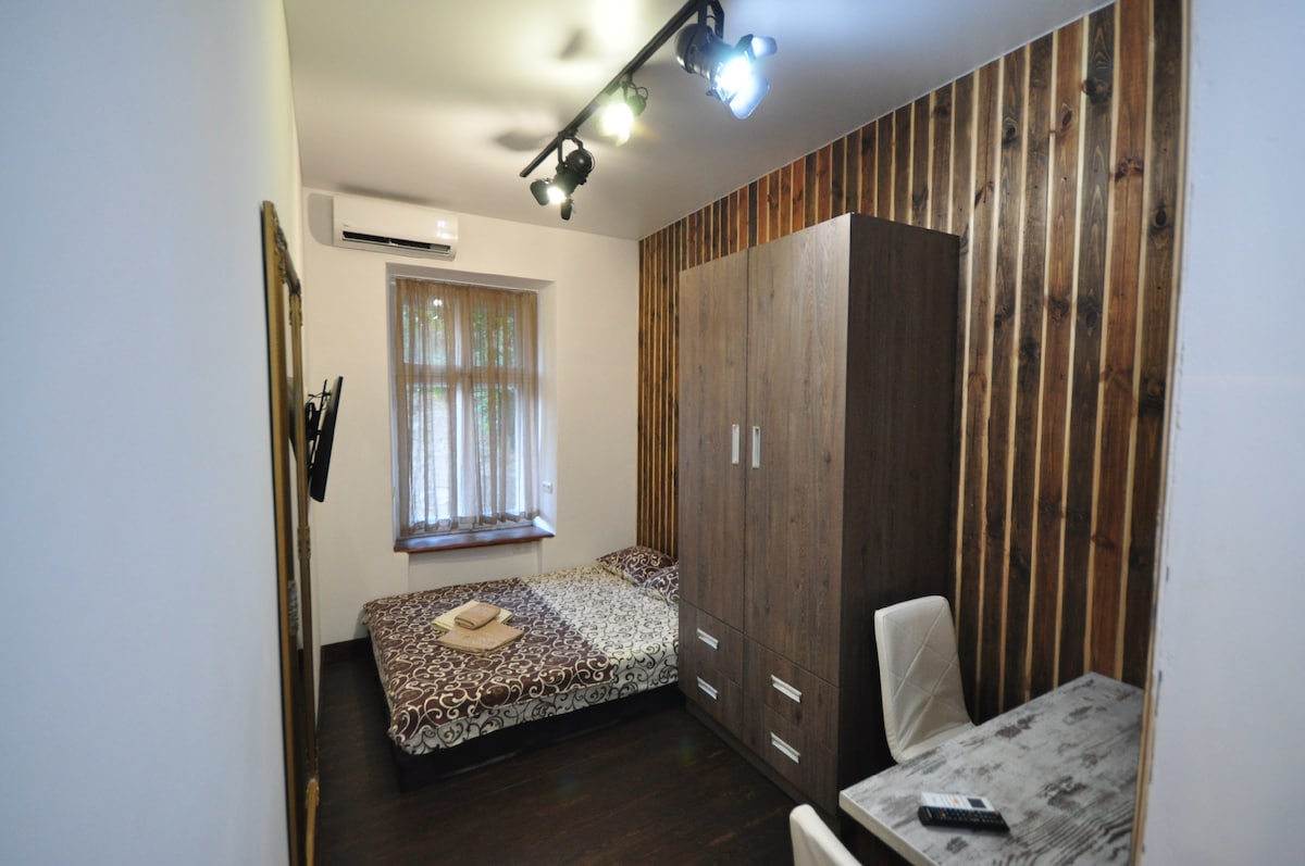 The studio apartment features a cozy sleeping area with a patterned bedspread covering a spacious bed. A wooden wardrobe is positioned against one wall, and a small desk with a chair is nearby. Natural light filters through the window, adding warmth to the interior.