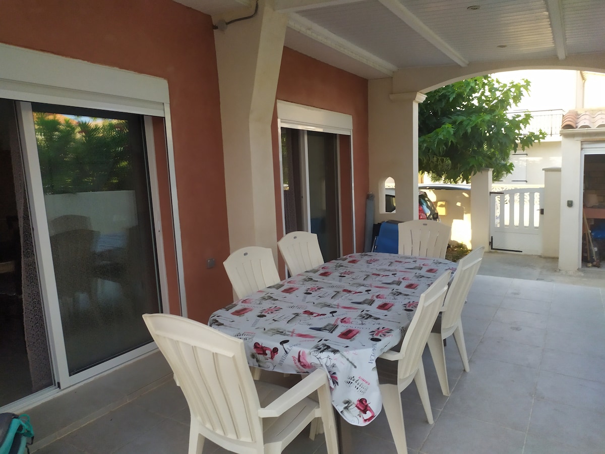 An outdoor terrace area features a large table set for dining, surrounded by six white chairs. Sliding glass doors provide access to the interior, allowing natural light to enhance the space. A leafy tree adds a touch of greenery nearby, contributing to the serene atmosphere.