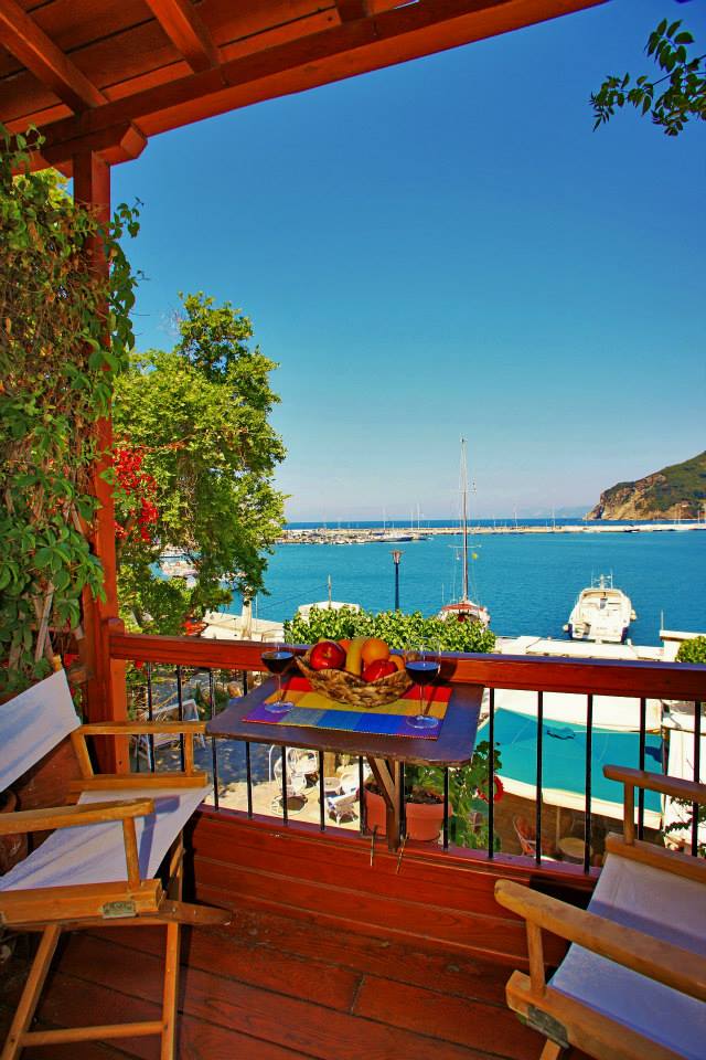 A charming balcony is surrounded by greenery and offers a view of the blue sea and harbor. A small table, set with fruits, and two wooden chairs are featured, inviting relaxation while enjoying the scenery. The bright sky enhances the tranquil atmosphere.