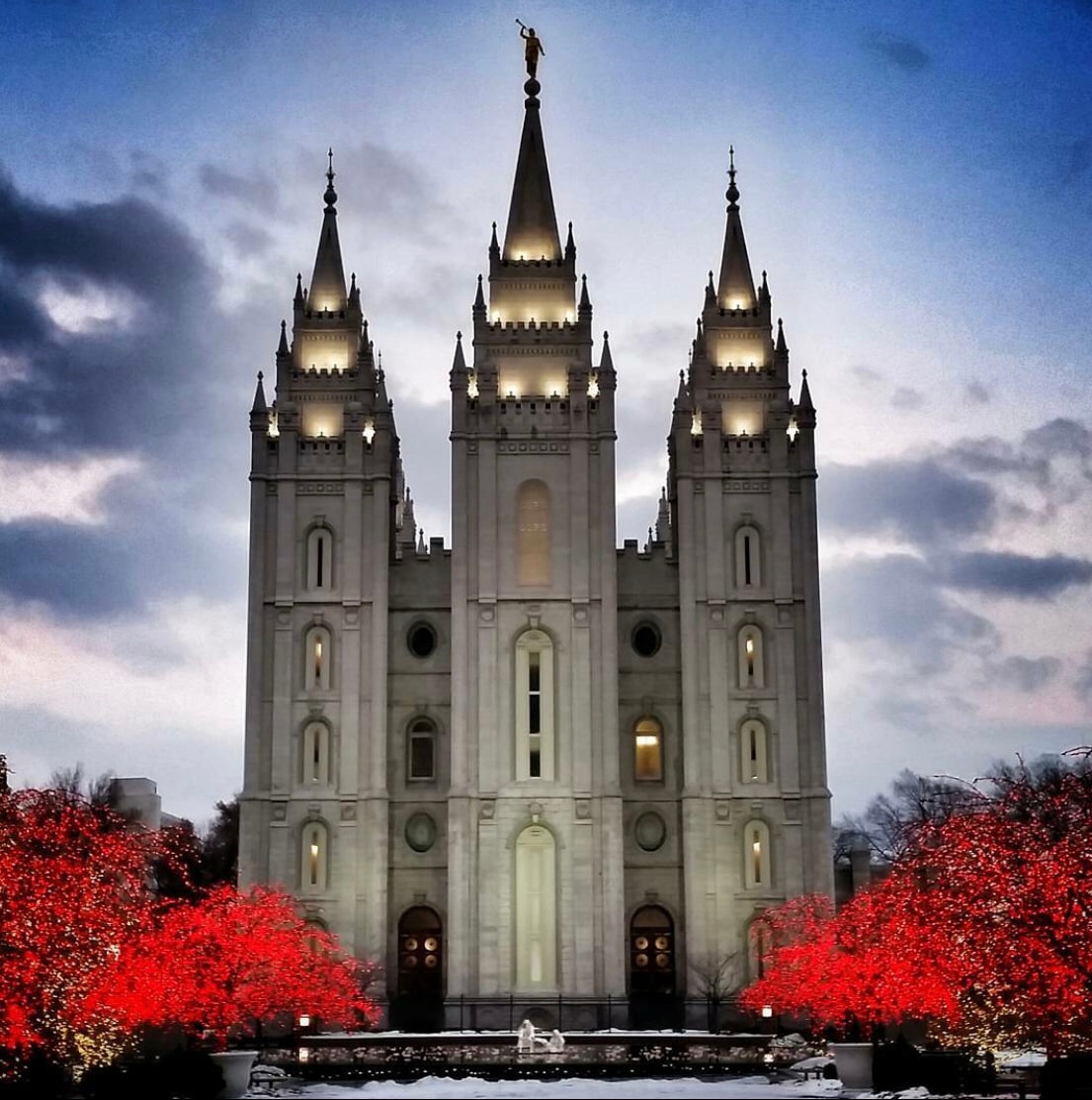 A striking architectural structure with tall spires is highlighted against a twilight sky. Bright red lights outline surrounding trees, complementing the building's illuminated features. The scene captures a serene winter atmosphere, enhanced by the snow-covered ground in front of the temple.