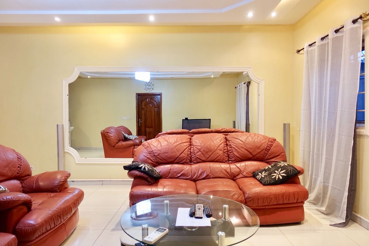 A spacious living area is highlighted by a large mirror that enhances the openness of the room. Comfortable red leather sofas are arranged around a glass coffee table, creating a relaxed setting. Soft white curtains frame the window, allowing natural light to filter into the space.