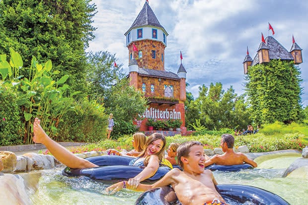 Guests can be seen floating in inner tubes on a lazy river surrounded by lush greenery. The iconic Schlitterbahn waterpark towers in the background, featuring distinctive turrets adorned with flags under a partly cloudy sky.
