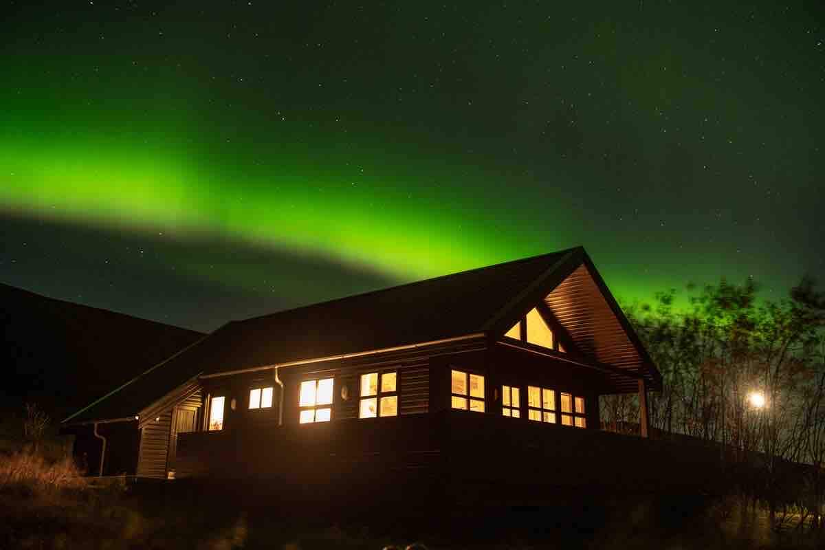The cabin, illuminated from within, is framed by a stunning display of the Northern Lights. Its large windows create a warm glow against the backdrop of the night sky, while the nearby landscape is graced with scattered trees and distant hills.